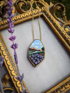 a necklace with a painting on it sitting in front of a frame next to purple flowers