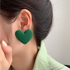 a close up of a person wearing green heart shaped earring with one hand on her ear