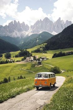 an old vw bus driving down a road in front of mountains and green grass