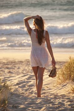 a woman walking on the beach with her hand in her hair and holding a purse