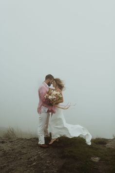 a bride and groom kissing on top of a hill in the foggy weather with their bouquet