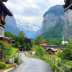 an empty road with mountains in the background and houses on both sides, surrounded by greenery