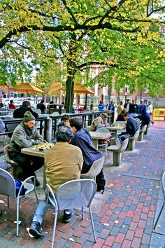 people are sitting at tables on the sidewalk in an open area with trees and benches