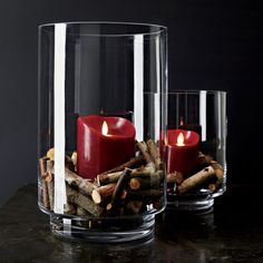 two glass vases filled with candles on top of a black table next to firewood