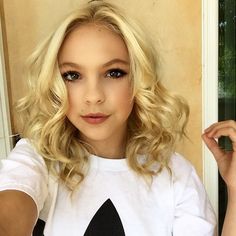 a woman with blonde hair wearing a white shirt and looking at the camera while standing in front of a door