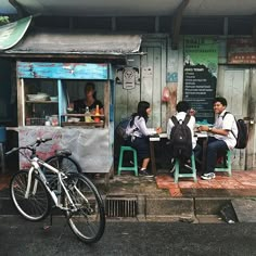 three people sitting at a table in front of a food stand with a bicycle parked next to it