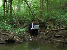 a man in a suit sitting on the edge of a river using a laptop