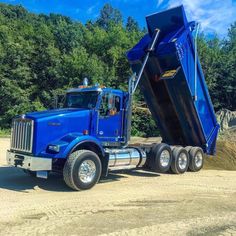 a large blue dump truck parked on top of a dirt road next to some trees