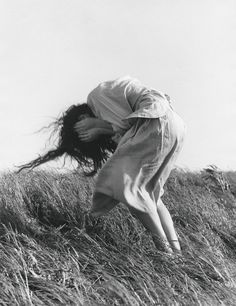 a woman jumping up into the air on top of a grass covered hill with her hair blowing in the wind