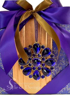 a purple and gold gift box with a ribbon around it's neck, decorated with an elaborate brooch