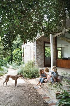 three children and a dog sitting on the steps in front of a brick house with trees