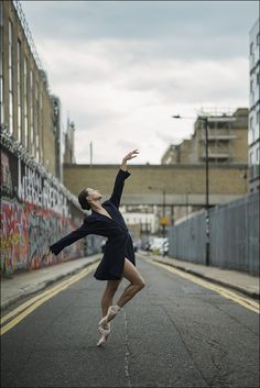 a woman in black dress dancing on street next to wall with graffiti painted on it