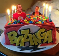 two men sitting in front of a birthday cake with candles on it and the word bazinga spelled out