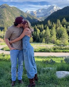 a man and woman hugging each other in the grass with mountains in the back ground