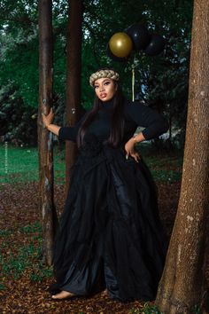 a woman in a long black dress standing next to a tree with balloons on it