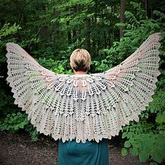 a woman is standing in the woods with her back turned to the camera and she has a crocheted shawl over her shoulders