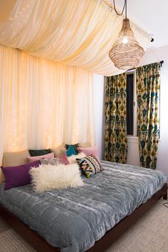 a bed with pillows and blankets in front of a curtained window that has lights hanging from the ceiling