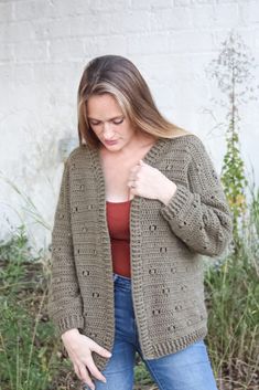 a woman wearing a green crochet cardigan and jeans standing in front of a white brick wall