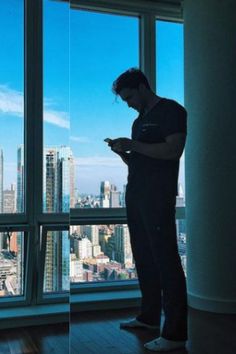 a man looking at his cell phone while standing in front of a large window overlooking the city