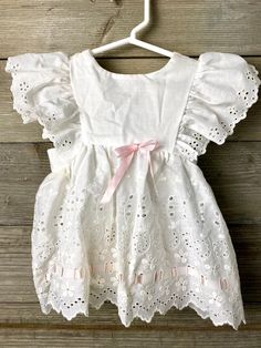 This is a white eyelet lace pinafore with pink ribbon accents. This is an apron style with a big bow in the back. This goes over a dress and is totally cottagecore! There is no size tag so I am estimating the size based on other listings I have. Please see the measurements for better fit.  Shoulders- 6" before the ruffles Waist- 10" flat across shoulder to bottom hem- 12" Pink Daywear Dresses With Bow, White Ribbon Dress For Dress-up, White Dress With Ribbon For Dress-up, Spring Baptism Dress With Broderie Anglaise, Summer Baptism Dress With Bow, Cute Baptism Dress With Lace Trim, Cute Baptism Dress With Lace Trim For Formal Occasion, Cute Lace Trim Baptism Dress For Formal Occasions, Fitted Broderie Anglaise Baptism Dress For Spring