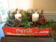 an old coca - cola crate is decorated with evergreen, pine cones and two candles