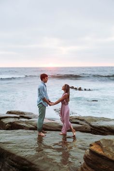 a man and woman holding hands while standing on rocks near the ocean