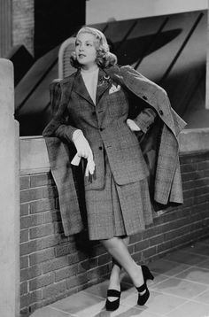 a black and white photo of a woman leaning against a brick wall wearing a coat