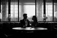 a man and woman sitting at a table talking to each other in front of windows