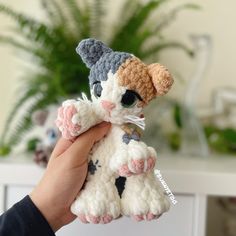 a hand holding a small crocheted cat toy in front of a potted plant