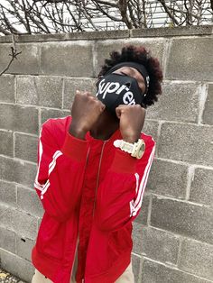 a young man wearing a red jacket and hat covering his face with one hand while standing in front of a brick wall