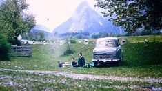 two people sitting on the grass next to an old car with mountains in the background