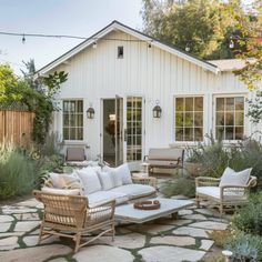 a patio with chairs, couches and tables in front of a white house surrounded by greenery