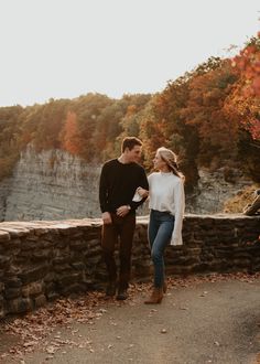 a man and woman standing next to each other near a stone wall with trees in the background