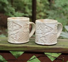 two coffee mugs sitting on top of a wooden table in front of some trees