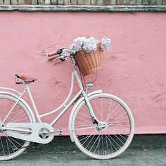 a white bicycle parked next to a pink wall with flowers in the basket on it