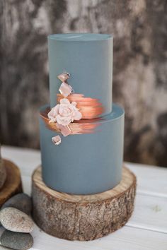 a three tiered blue cake with pink flowers on the top and bottom, sitting on a tree stump