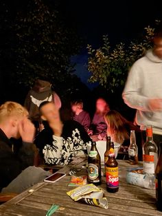 a group of people sitting around a wooden table with beer bottles and snacks on it