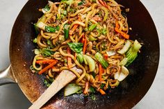 a wok filled with noodles and vegetables on top of a white countertop next to a wooden spoon