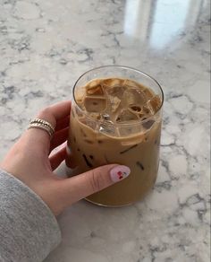 a hand holding a drink with ice in it on a marble counter top next to a woman's finger