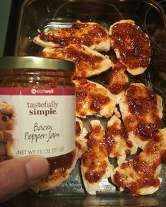 a person holding a jar of jam next to some chicken wings in a tin on a table