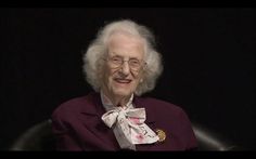 an old woman with glasses and a bow tie sitting in a chair smiling at the camera