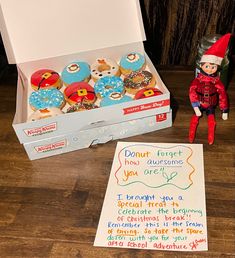 an elf is sitting next to a box of donuts and a handwritten note