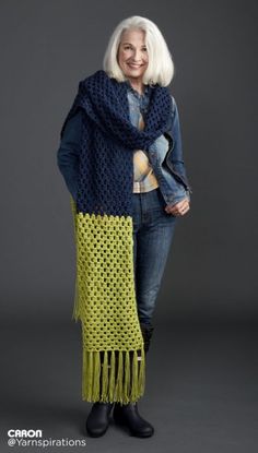 a woman with white hair wearing a blue and yellow knitted shawl, standing in front of a gray background