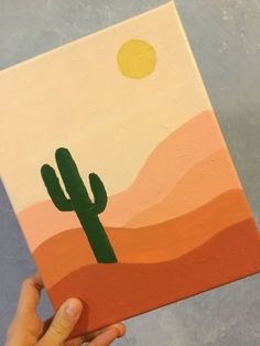 a person holding up a painting with a cactus on it's side and the sun in the background