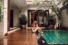 a woman sitting on the edge of a pool in front of a house with plants