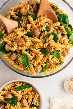 a bowl filled with pasta and spinach next to two wooden spoons on a white surface