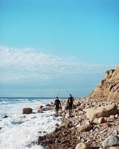 two people standing on rocks near the ocean with fishing poles in their hands and one person holding a surfboard