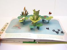 an open book with paper cutouts of plants and animals on it, sitting on a table