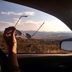 a person holding up their sunglasses in the back seat of a car with mountains in the background