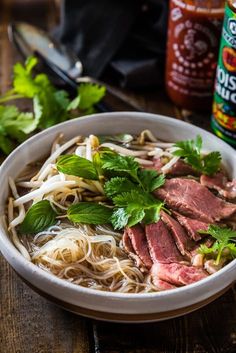 a white bowl filled with meat and noodles next to a jar of chili sauce on top of a wooden table